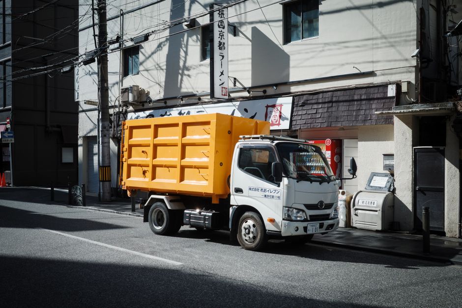 A small commercial waste collection truck with a white cab and a bright yellow, textured, metal dump body is parked on a city street in front of a building with traditional Japanese signage. The truck