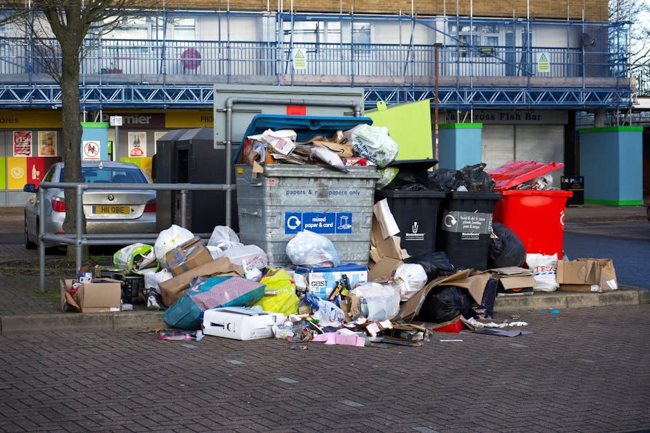 A large pile of mixed household rubbish and discarded packaging materials is overflowing from a grey wheeled bin labeled for mixed paper and cardboard, situated on a paved area beside a curb. Surround