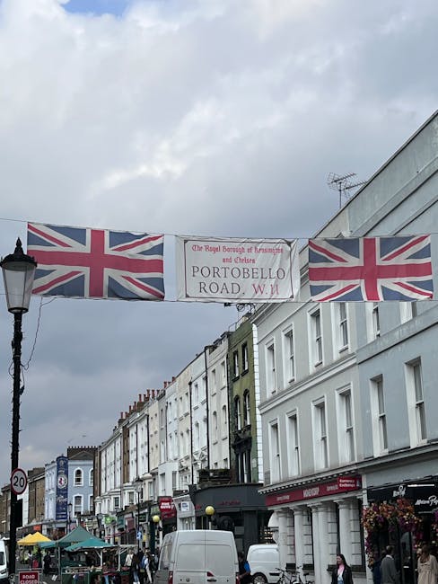 A busy street scene on Portobello Road featuring rows of multi-story Victorian-style buildings with detailed facades and large windows, some with decorative molding and balconies. The street is filled